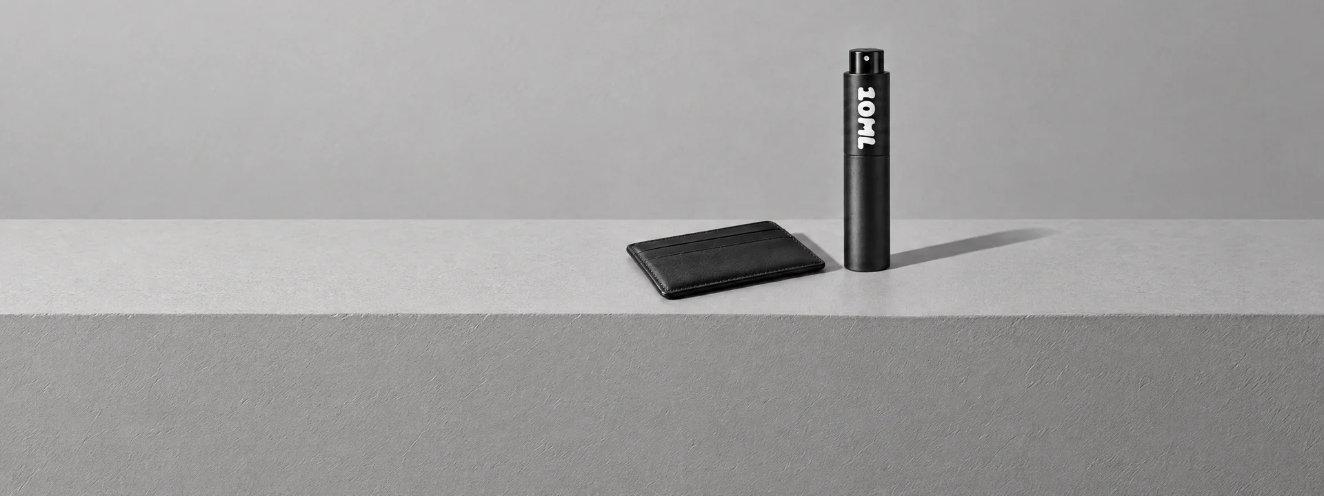 A black 10ML atomiser beside a leather card holder on a grey plinth