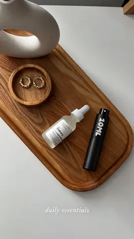 A black 10ML atomiser on a wooden tray with gold earrings and a serum, captioned “daily essentials”